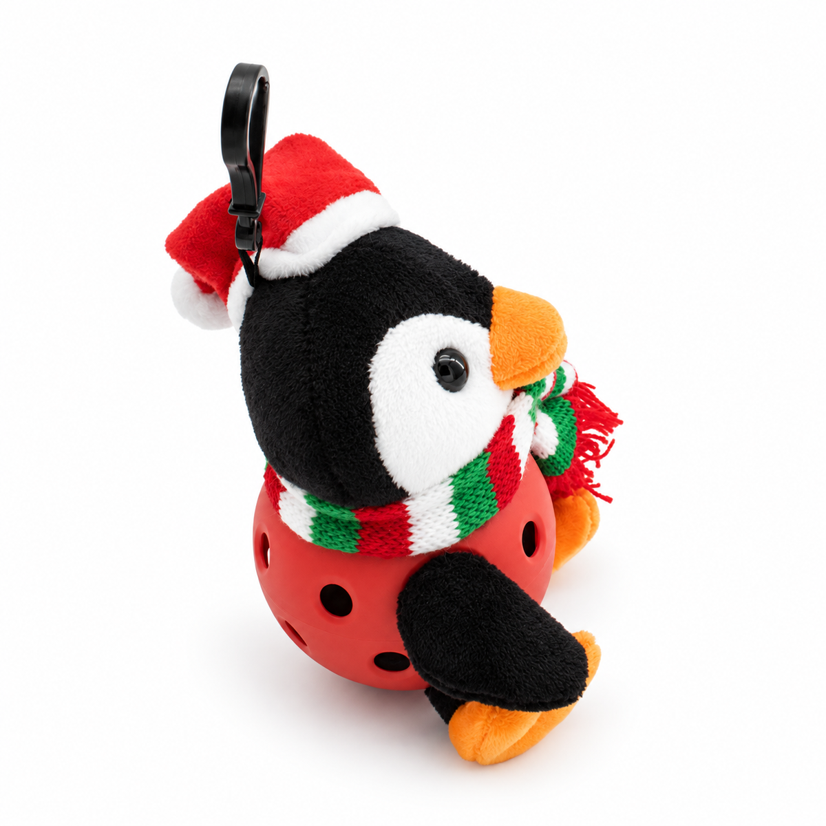 A plush penguin keychain or bag charm wearing a red Santa hat with white trim and a red, green, and white striped knit scarf, attached to a black plastic carabiner clip. The penguin is in a red pickle ball and orange feet and beak, set against a plain white background.