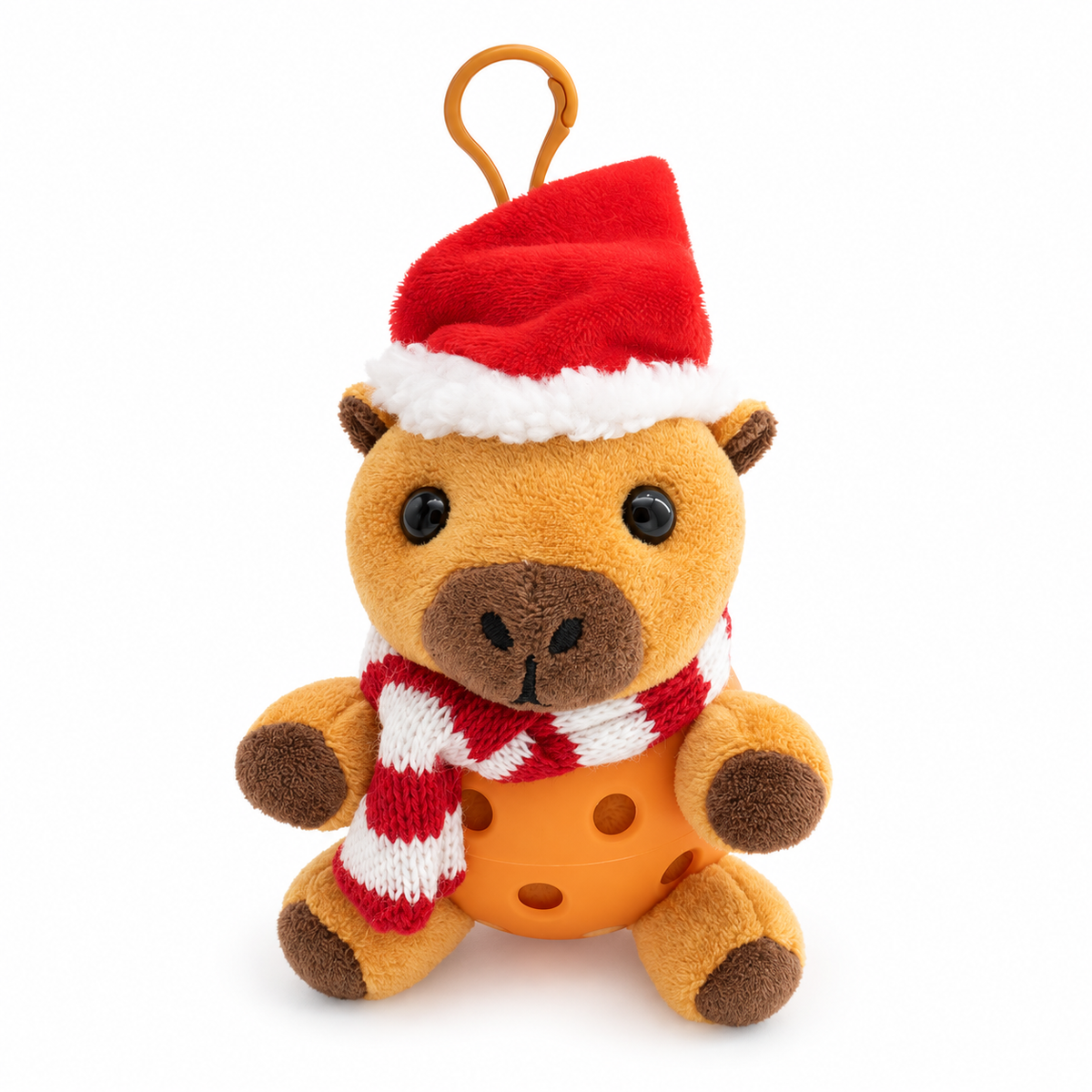 A small plush capybara with a Santa hat and striped red-and-white scarf, sitting in an orange pickle ball against a clean white background.