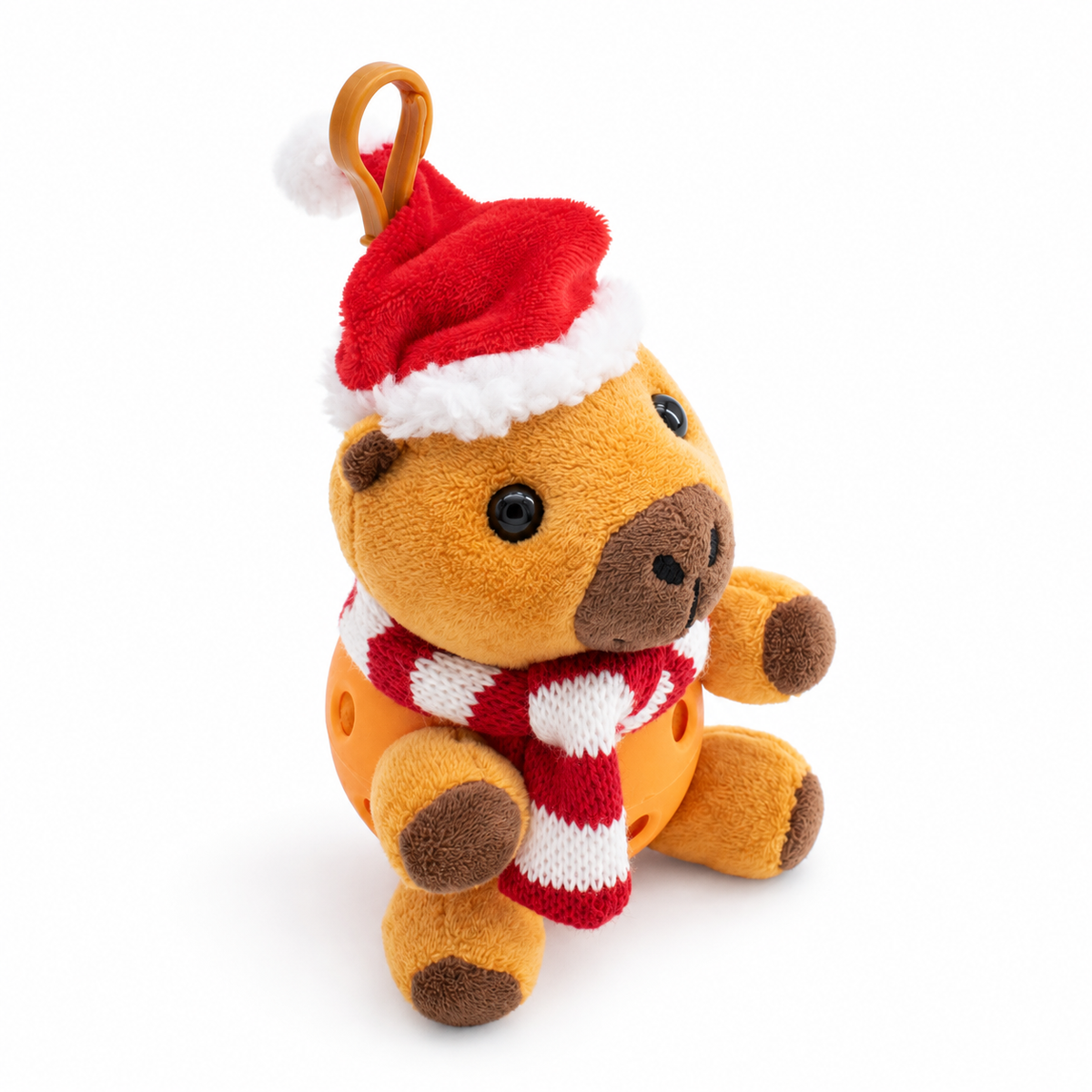 A small plush capybara with a Santa hat and striped red-and-white scarf, sitting in an orange pickle ball against a clean white background.