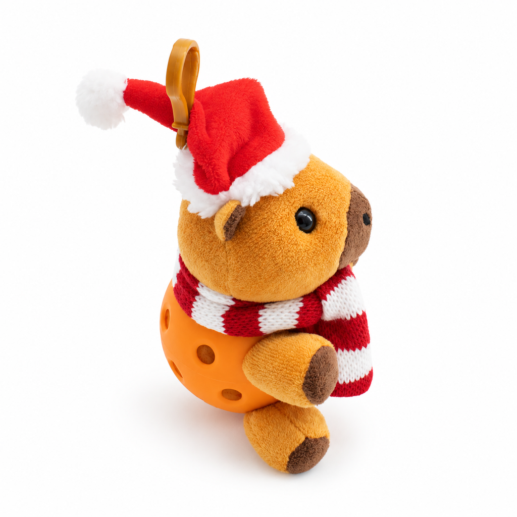 A small plush capybara with a Santa hat and striped red-and-white scarf, sitting in an orange pickle ball against a clean white background.